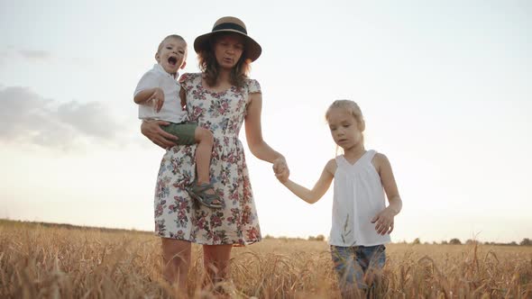 A Mother in a Summer Dress and Hat with Her Son and Daughter Walks Through a Wheat Field