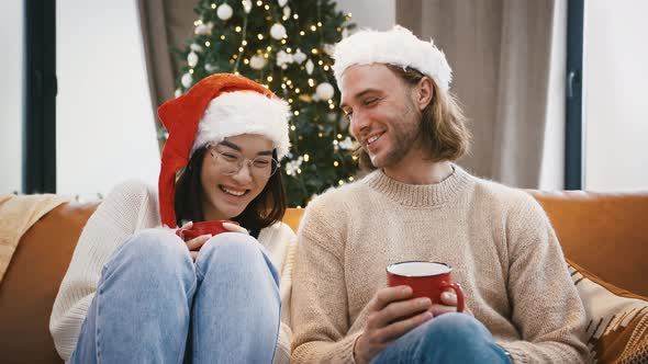 Multiracial Male and Female Laughing Talking Holding Mugs of Tea Sitting on Couch Against Decorated