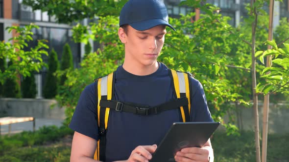 Portrait Food Delivery Young Man Courier with Thermal Backpack Uses Tablet