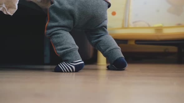 Funny Baby in Grey Pants Plays at Bed on Wooden Floor