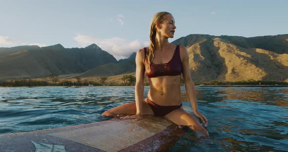 Young woman sitting on a surfboard at sunset alt