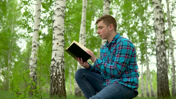 A Young Man Is Sitting in Nature with a Book in His Hands. A Man Sits on a Stump in a Birch Forest