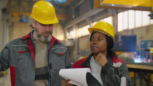 Diverse Coworkers in Hardhats Walking and Speaking in Machinery Plant