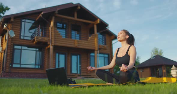 Young Woman Meditates with Laptop in the Backyard on the Green Grass Against the Background of