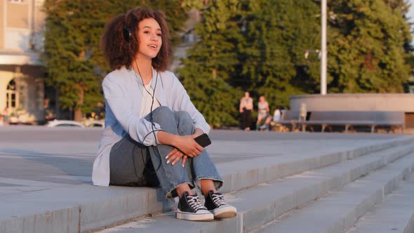 Young Afro American Girl Student African Woman Sitting on Street Outdoors in City Wears Headphones