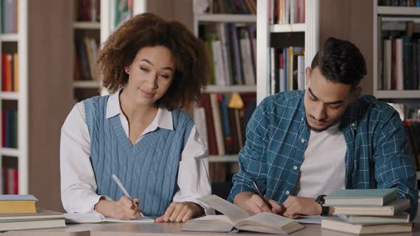 Two Students Young Guy and Girl Doing Homework Sitting at Desk in Library Writing Notes Attentively