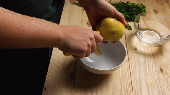 Women's hands peel potatoes with a potato peeler. The peel from the vegetable falls into a cup