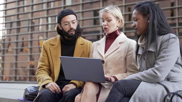 International Business Coworkers Discussing Project on Laptop Sitting Together on Bench Outdoors in