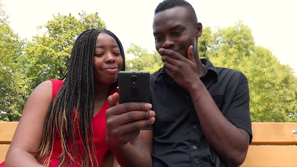 A Black Man and a Black Woman Sit on a Bench in a Park, Look at a Smartphone and Laugh