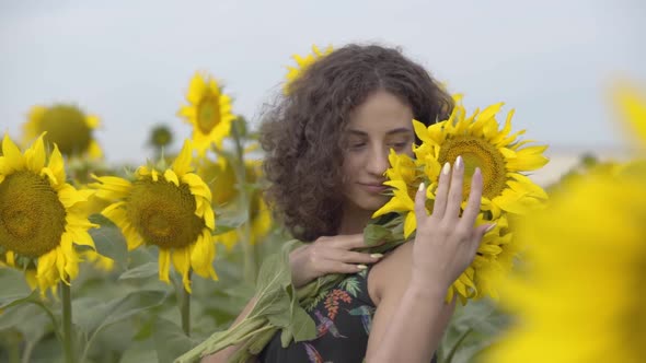 Portrait of a Beautiful Curly Girl Standing on the Sunflower Field with the Bouquet of Flowers