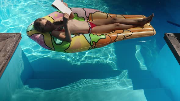 Happy Smart Girl Sunbathing on Inflatable Mattress in Swimming Pool Reading Book in Sunbeam