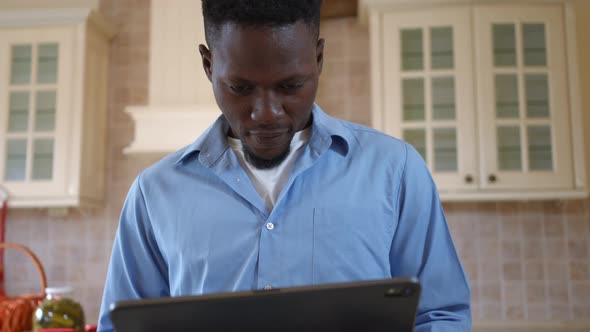 Front View Smiling Young Man Messaging Online on Tablet Standing in Kitchen at Home