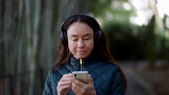 Middleaged Woman is Listening to Favorite Songs By Wireless Headphones and Cell Phone During Walk