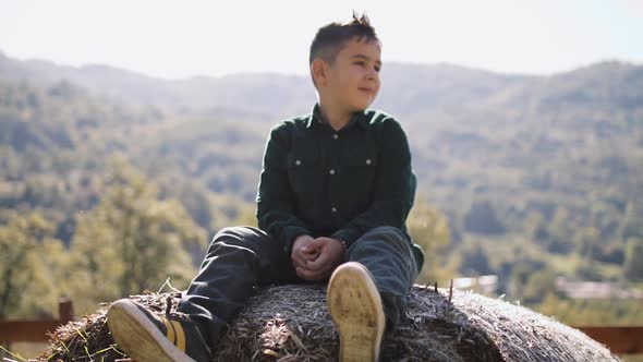 A Little Boy of 8 Years Sits on a Haystack and Looks at the Mountains