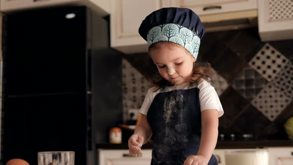 A Little Funny Girl in a Chef's Hat Eats Flour with Her Hands on the Kitchen.