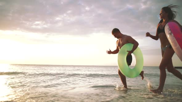 Happy Young Couple Runnung on the Beach with Rubber Rings During Sunset Slow Motion