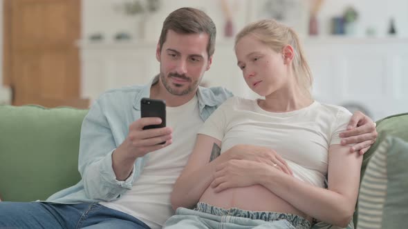 Pregnant Couple Using Smartphone While Sitting on Sofa