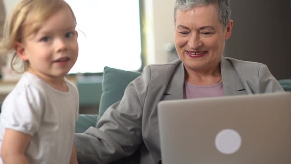 Portrait of a Stylish Grandmother Working at Home on the Couch and Her Little Blondie Grandson