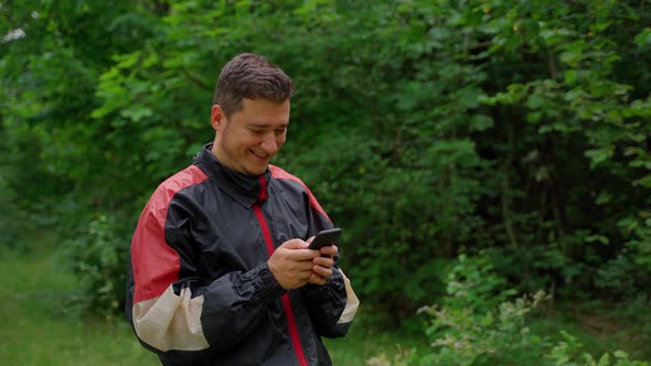 30s man holding smartphone smile in forest. Attractive young adult male person using telephone