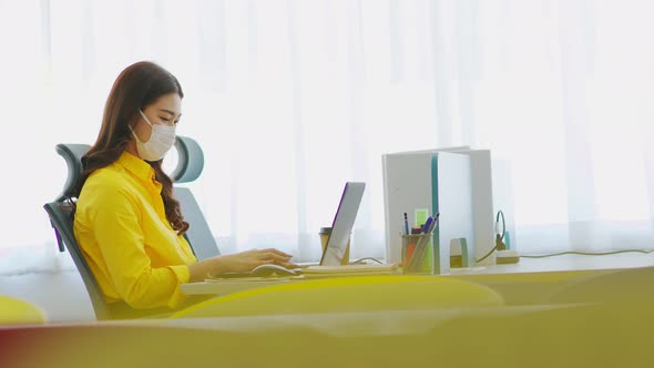New normal of Asian woman in yellow shirt wearing surgical face mask working with computer laptop