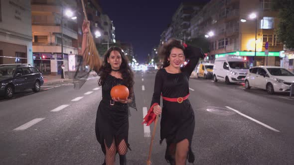 Girls In Witch Costumes Walking At The Road And Holding Pumpkin And Broomsticks