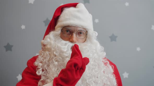 Portrait of Santa Making Shush Hand Gesture and Looking at Camera on Grey Starry Background