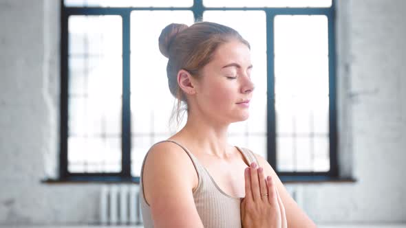 Calm young woman with hair bun in beige top does yoga breathing exercises holding hands in namaste