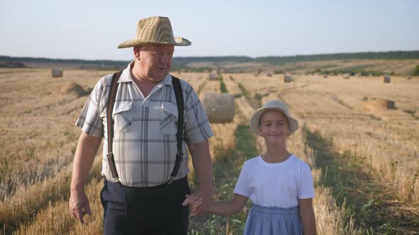 Grandfather and Granddaughter Walking Across the Field with Haystacks. Farmer Grandfather Teaches