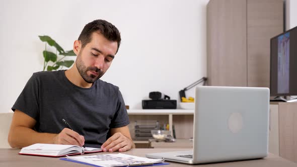 Businessman at Home in Casual Clothes with a Laptop on the Desk Takes Notes in a Notebook