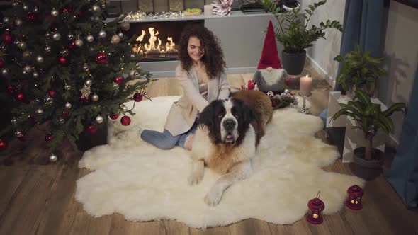 Smiling Brunette Caucasian Girl Sitting with Big Dog on Soft Carpet in Front of Fireplace. Decorated