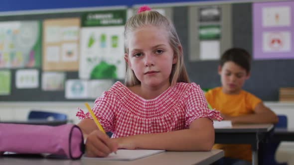 Portrait of happy caucasian schoolgirl sitting at classroom, making notes, looking at camera