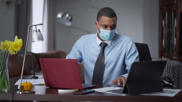 Portrait of Overworking African American Man in Covid Face Mask Typing on Laptop and Tablet