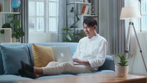 Smiling Asian Man Having Video Call On Laptop Computer While Lying On Sofa In The Living Room