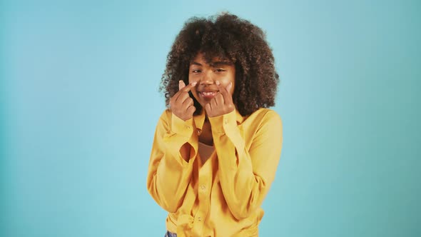 Young Darkskinned Female Laughing and Showing Finger Heart Hand Gesture While Posing Against Blue