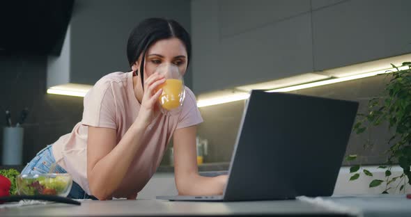 Brunette Working on Laptop which Standing on Kitchen Table and Drinking Orange Juice