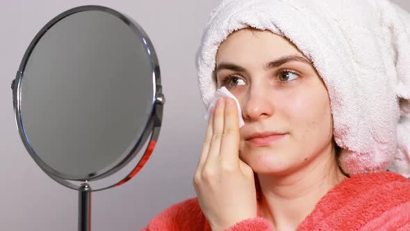 A Young Woman in a Pink Robe Applies Tonic to Her Face and Neck Using a Cotton Pad