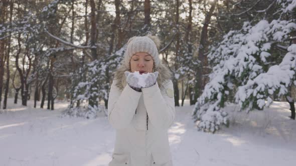 Happy Cute Girl Dressed in Warm Winter Clothes Blows Off Snow From Her Hands to Camera Outdoors in