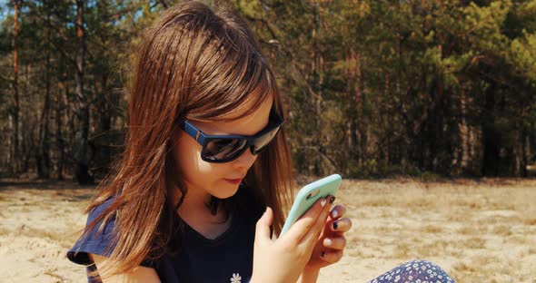 Cute Little Girl Holds Phone in Hand and Sits in Summer Forest
