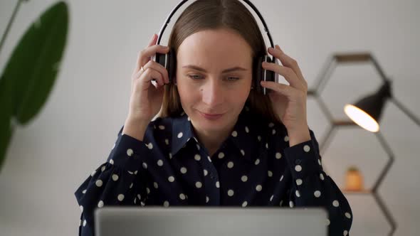 A Young Female Project Manager Puts on Headphones and Works on a Laptop Computer in an Office