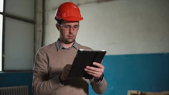 The Man Holds a Tablet in His Hand, on the Head of a Builder a Protective Helmet