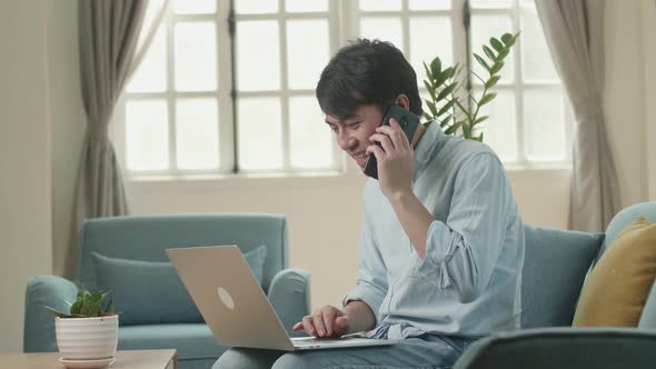 Asian Man Sitting Sofa In Living Room While Speaking By Phone And Working On Laptop