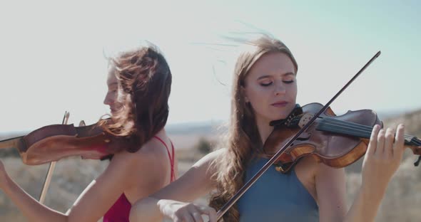 Close View of Female Violinists Duet in Blowing Dresses Play Among Cliffs