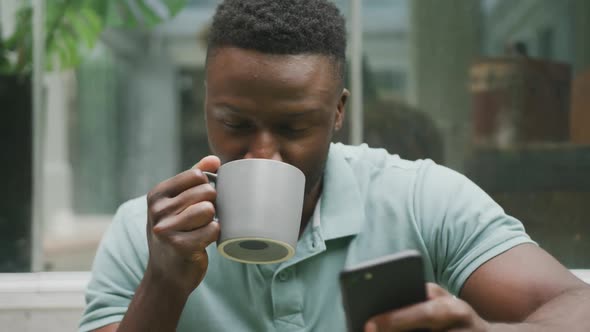 Happy african american man sitting with smartphone and drinking coffee in garden