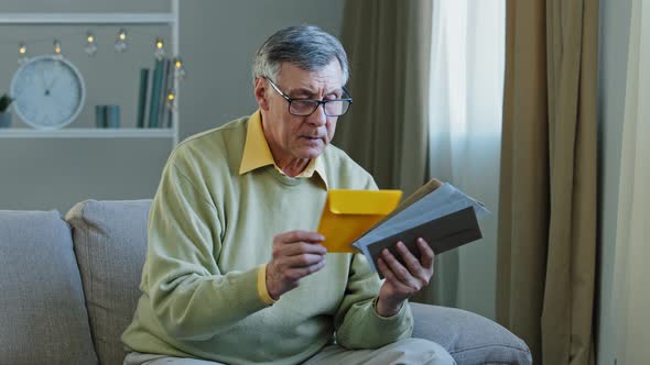 60s Elderly Man Recipient with Glasses Sorting Letters in Living Room Concentrated Mature