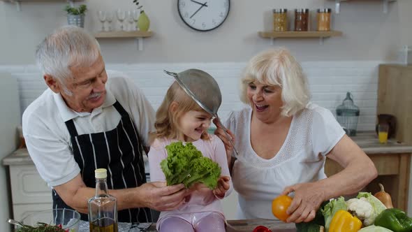 Senior Woman and Man with Grandchild Girl Making a Funny Dance with Strainer and Vegetables at Home