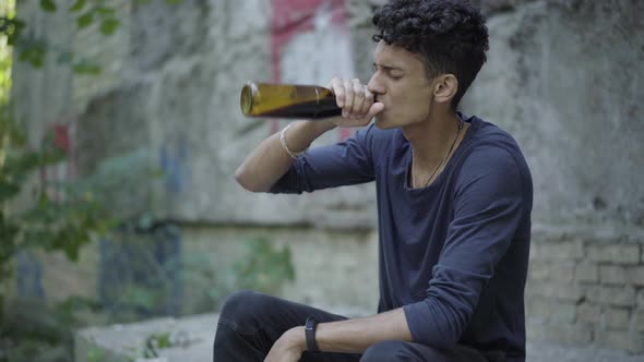 Frustrated Teenage Boy Drinking Beer at Slum and Crying. Side View Portrait of Depressed Mixed-race