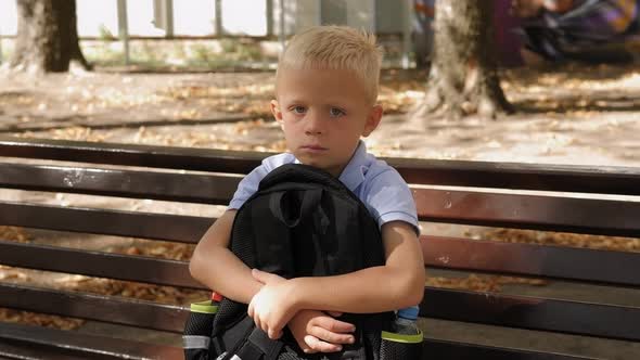 Sad Little Boy Sitting Alone on a Bench and Holding His School Backpack