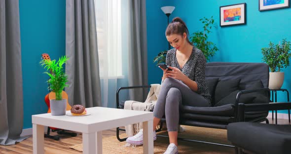 A Young Woman is Relaxing on the Living Room Couch Texting on Her Smartphone