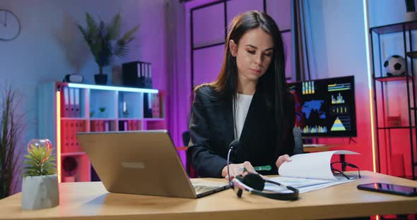 Businesswoman Working with Information on Laptop and Browsing Paper Report in Evening Office