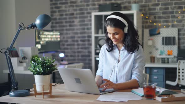 Attractive Lady in Headphones Listening To Music at Work and Working with Laptop